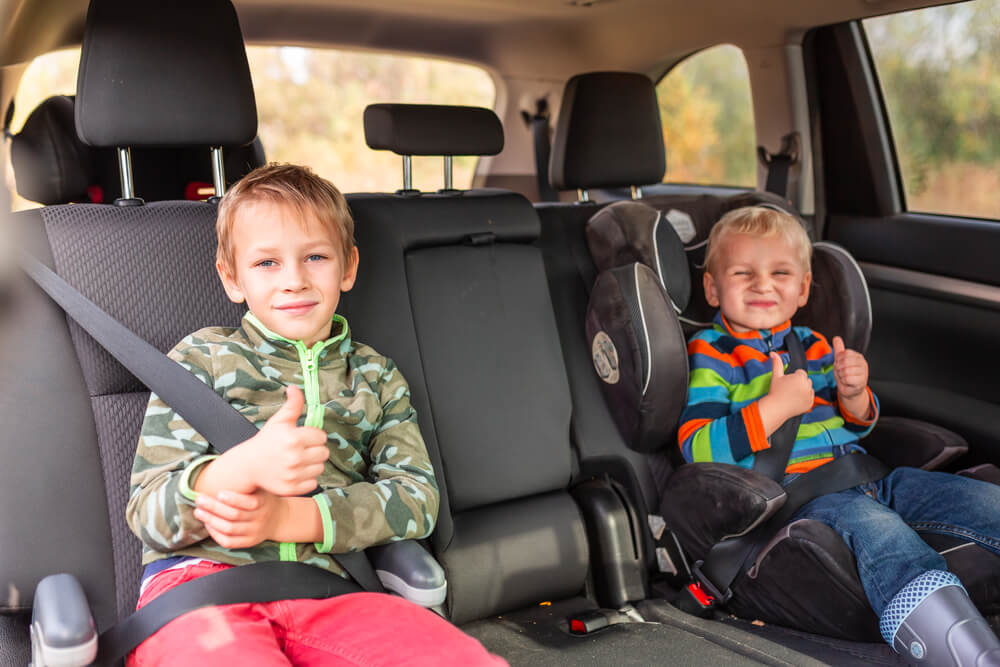 Two little boys sitting on a car seat and a booster seat buckled up in the car.